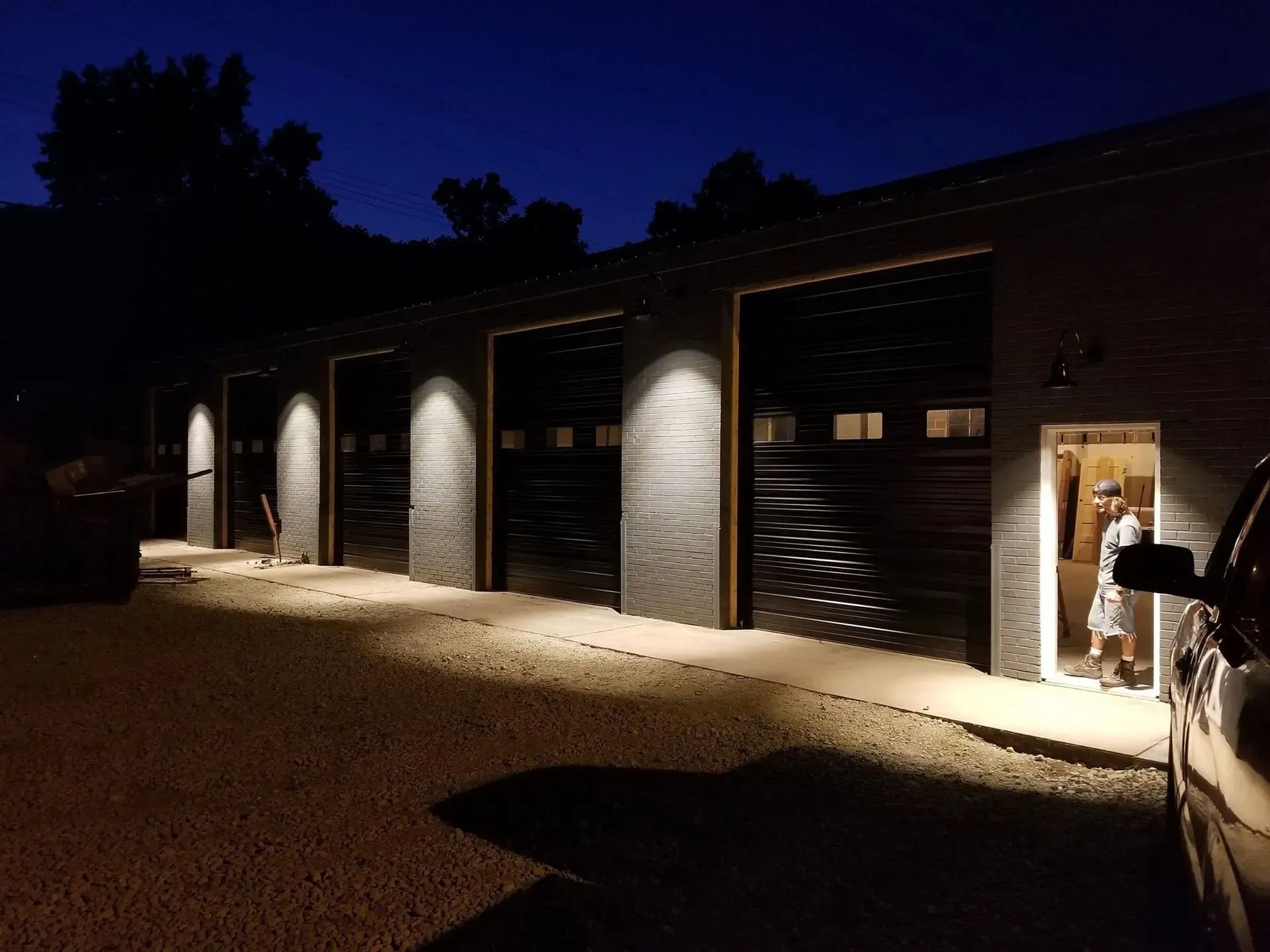 Dark exterior of a garage with open bays, illuminated at night. Person in doorway.