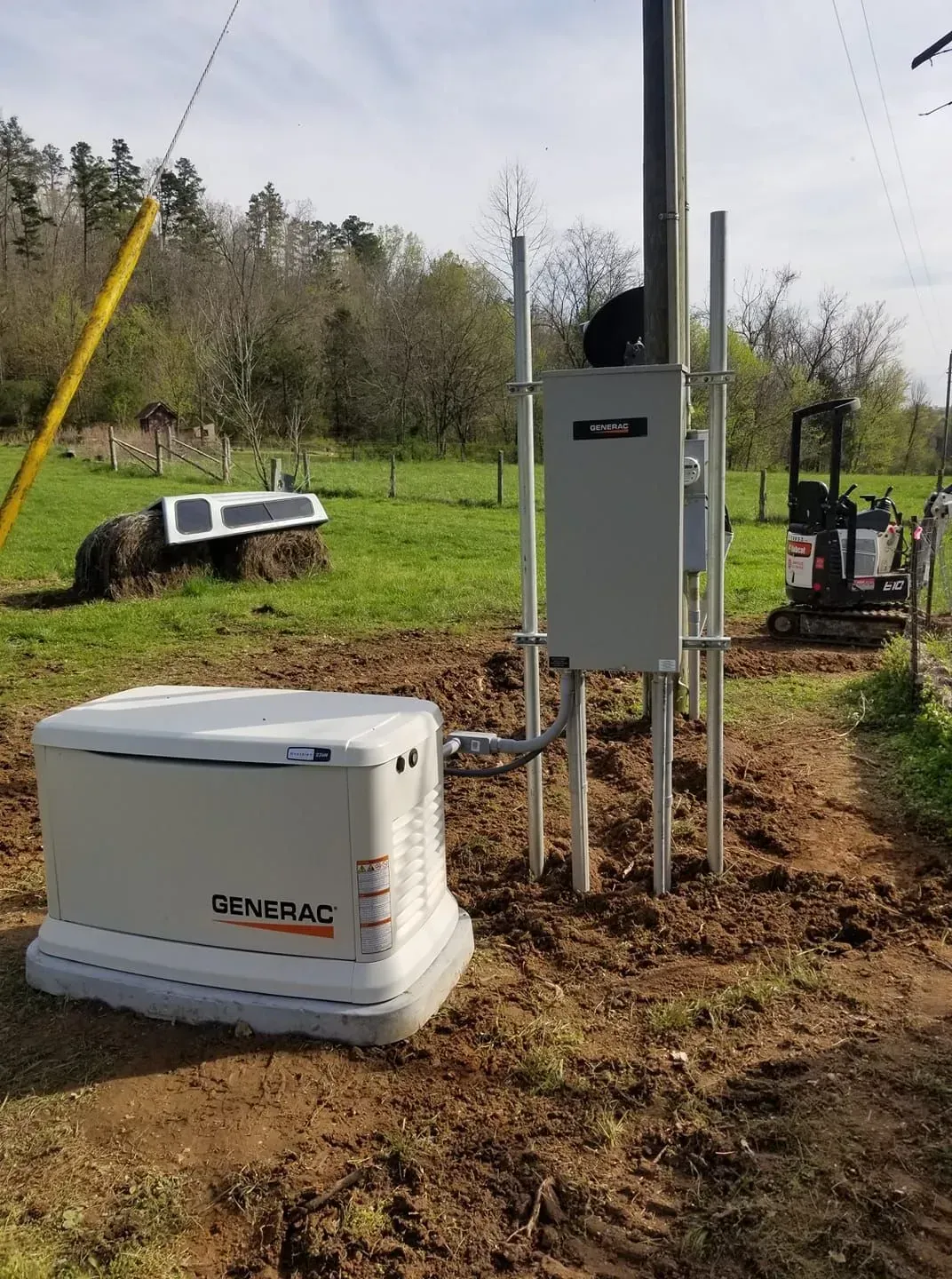 A Generac generator installed next to a utility pole in a grassy field, with an excavator in the background.