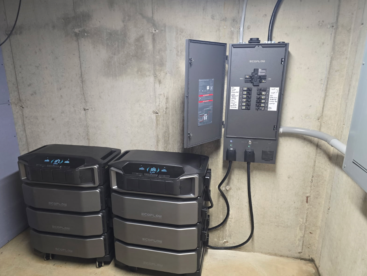 Two stacked battery units and electrical panel on a concrete wall in a basement.