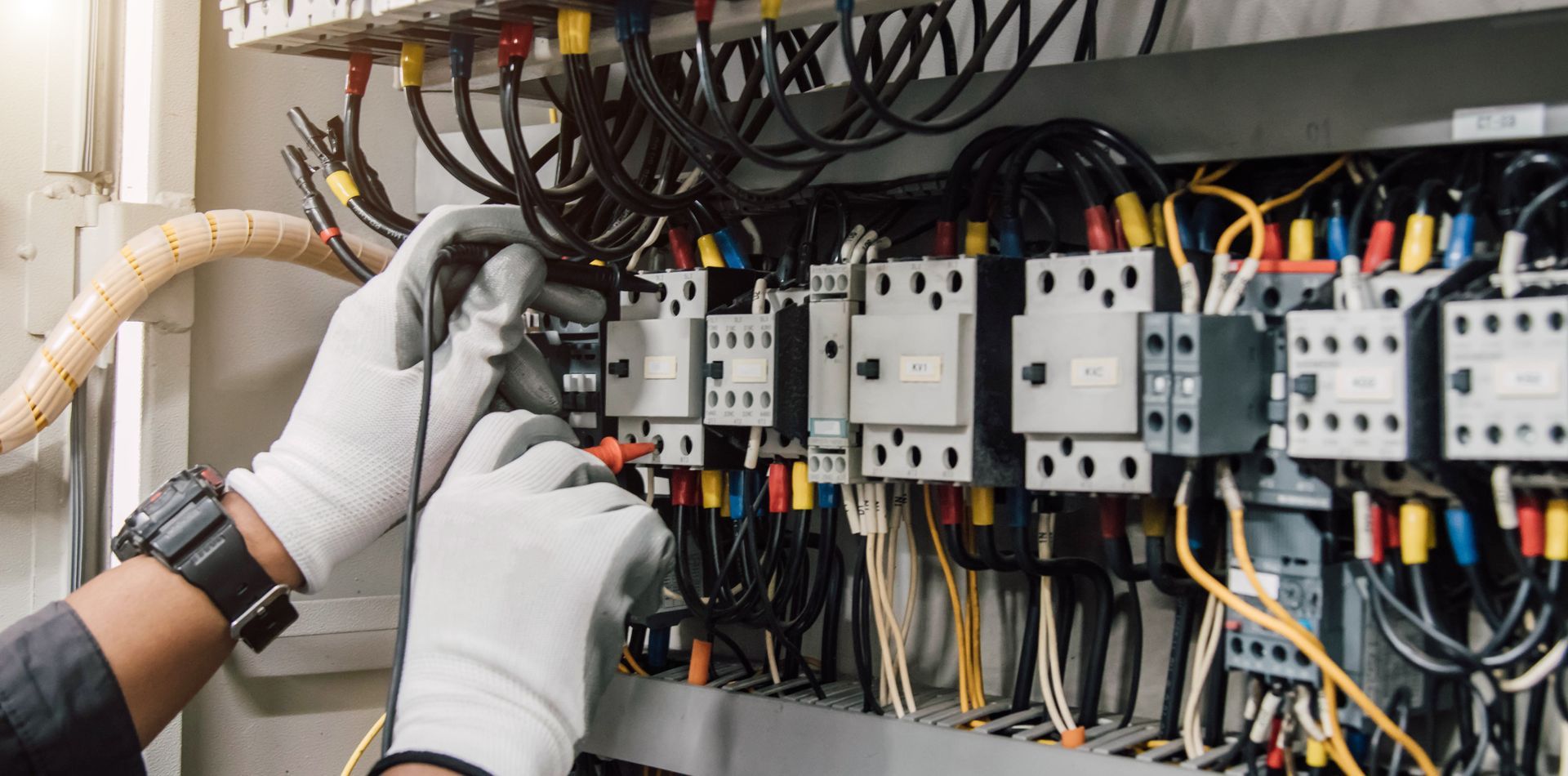 An electrician with white gloves is working on wiring in a panel box. Wires are various colors.