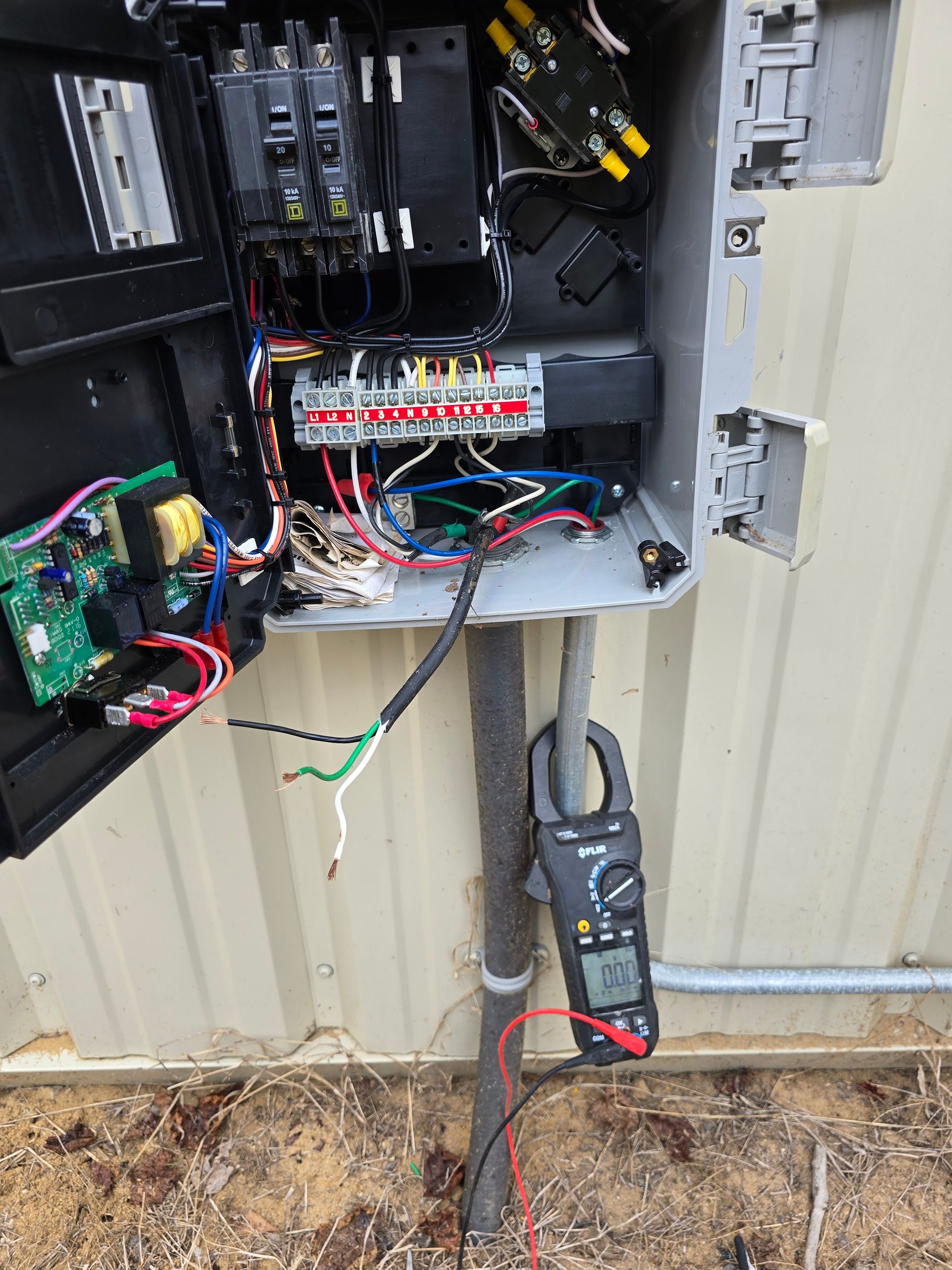 Open electrical panel with wires and a multimeter, mounted outdoors.