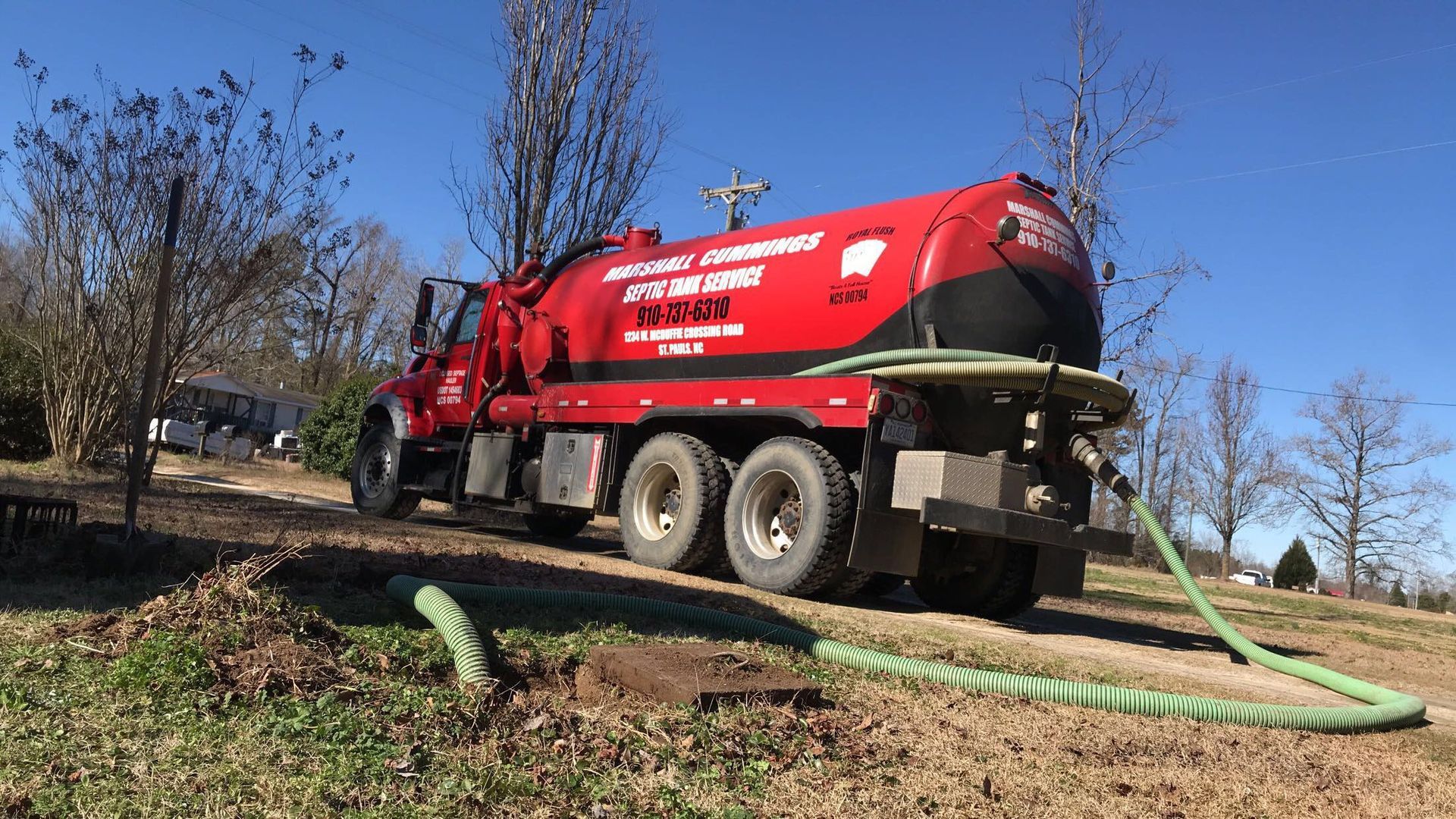 Red septic truck parked on a grassy hill; green hose extends to the ground, trees in the background.