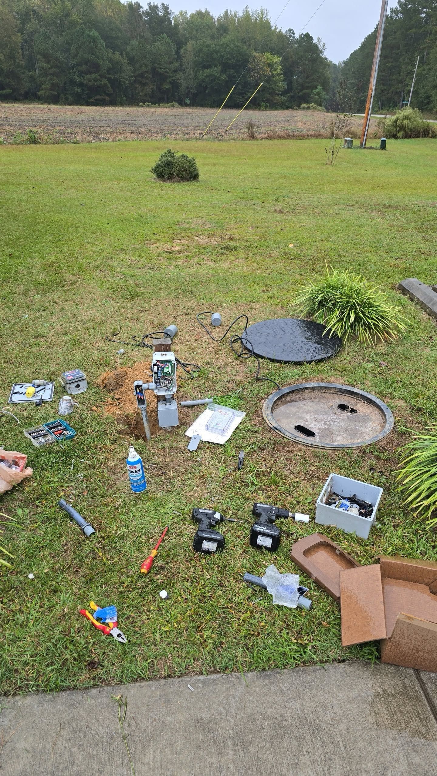 Tools and parts scattered on wet grass near two open sewer access covers; overcast day.