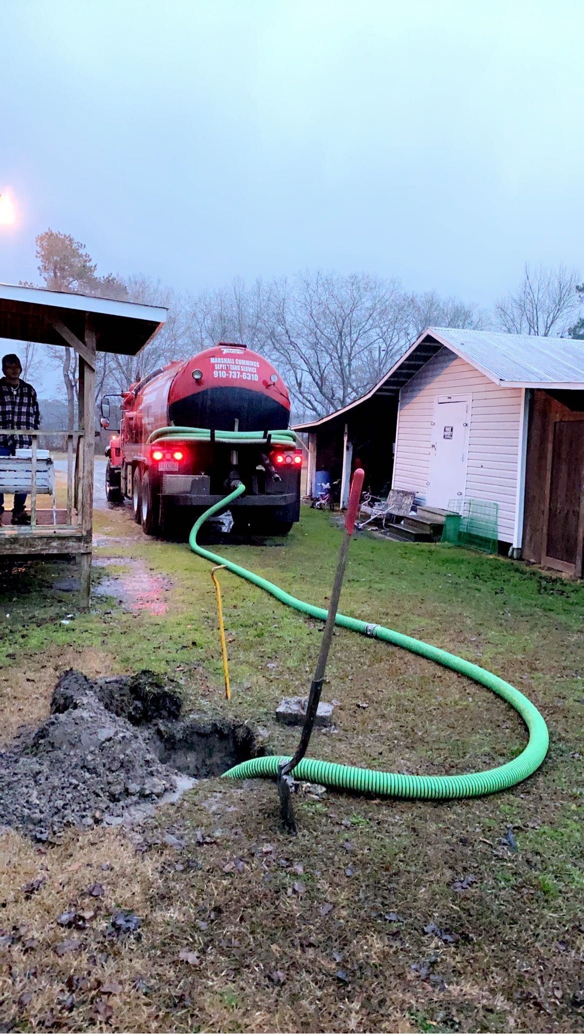 Septic tank truck pumping a system in a rural yard; green hose in ground, person on porch, snow-dusted trees.