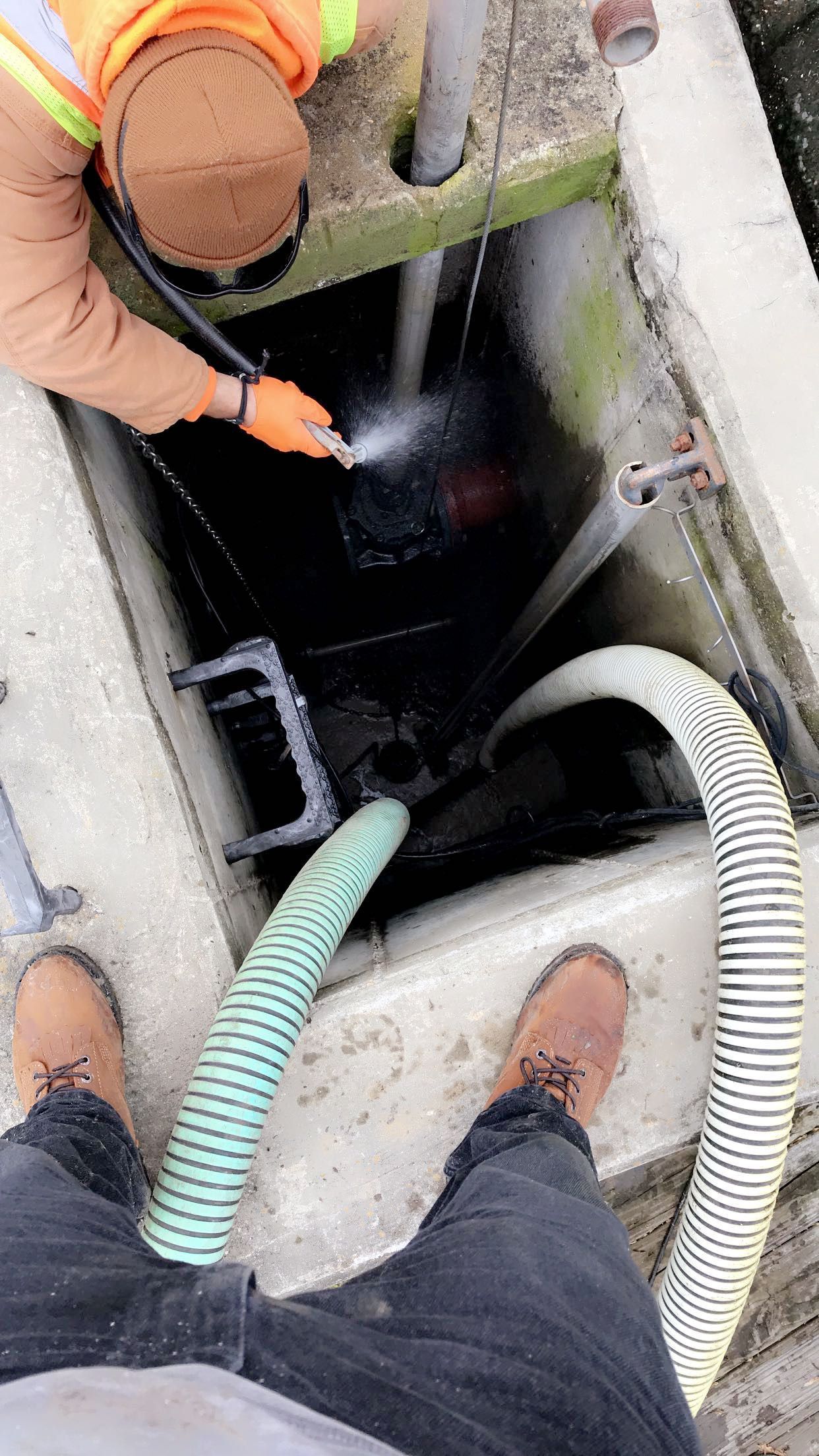 Person cleaning a dark industrial drain with a hose. Two hoses feed in, person wears a reflective vest and gloves.