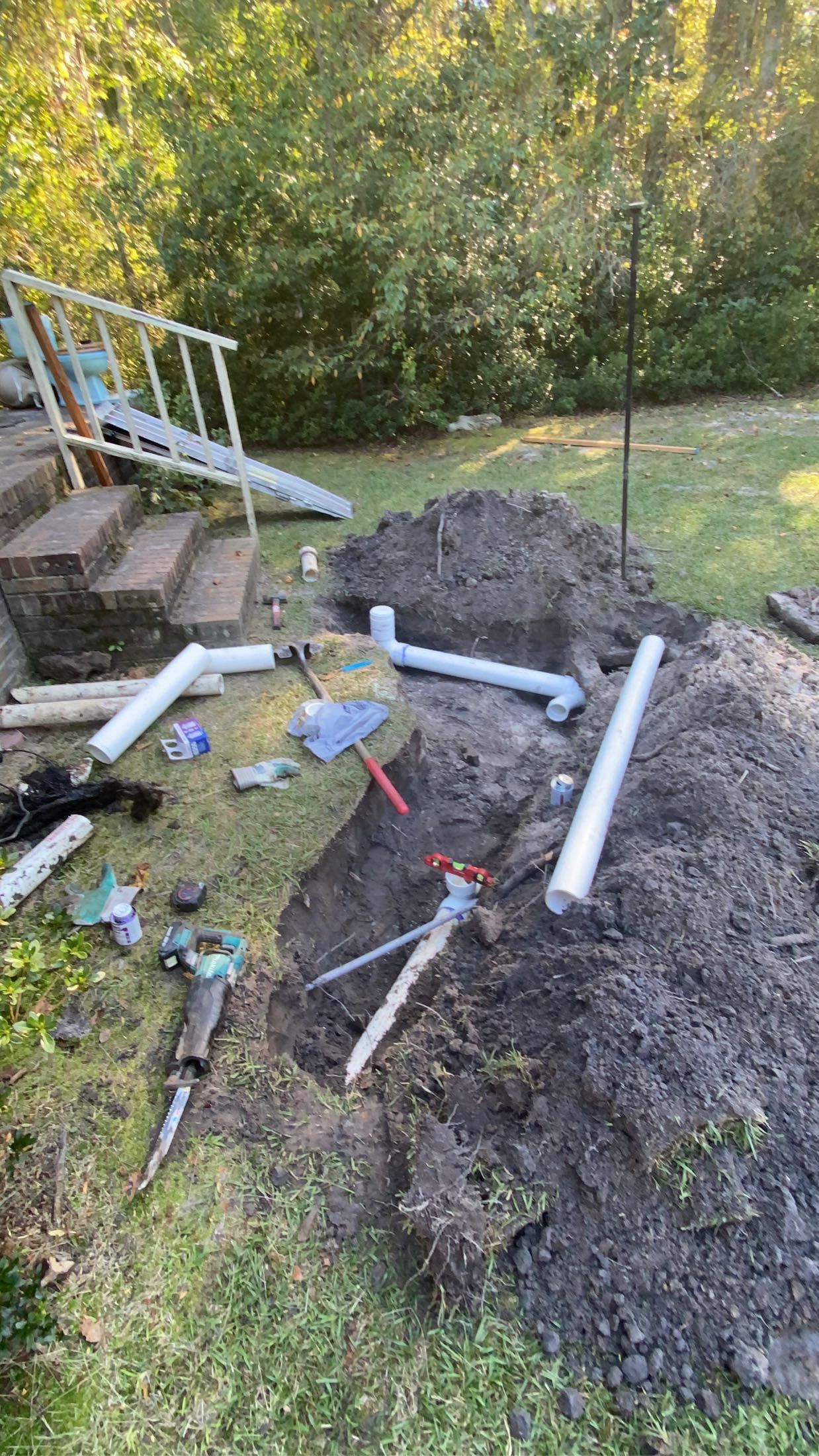 Outdoor scene of dug-up ground with plumbing pipes and tools scattered. A set of stairs is in the background.