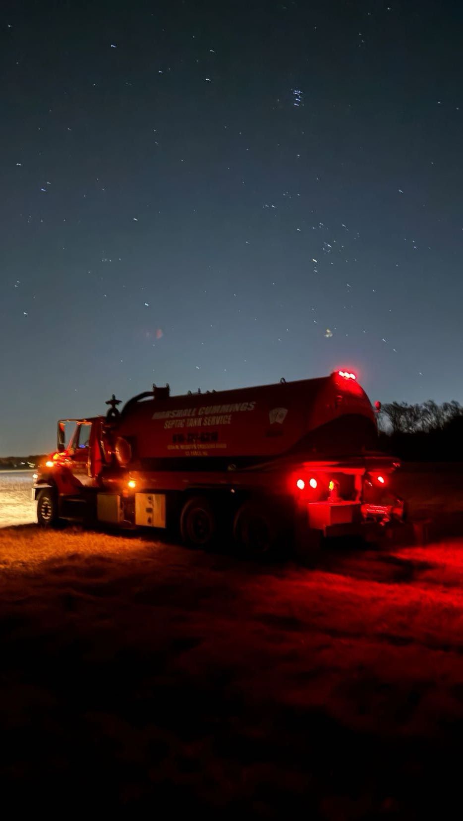 Red tanker truck parked under a dark night sky with visible stars; red tail lights and headlights illuminate the scene.