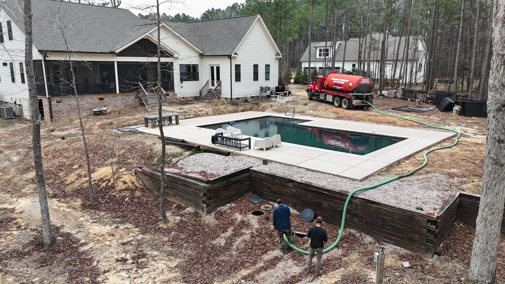 Pool being filled, brickwork in foreground, red tanker truck in background, two people near hose.