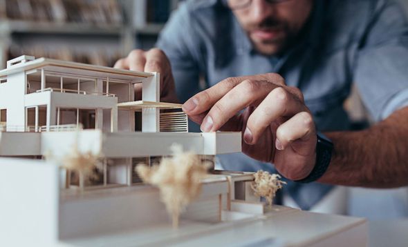 Hands adjusting a small architectural model on a table, with a blurred person in the background