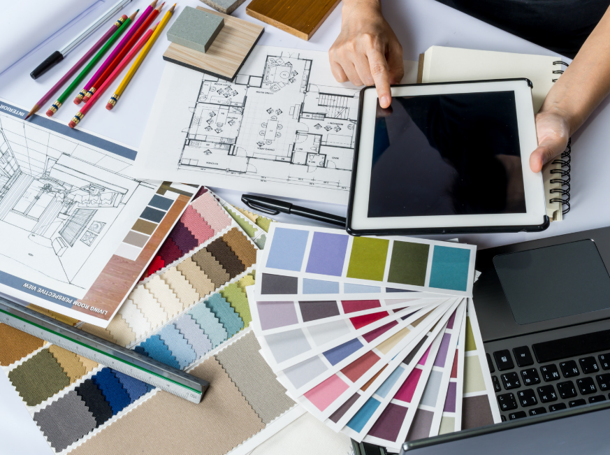 Interior design workspace with floor plans, color swatches, pencils, and a hand holding a tablet