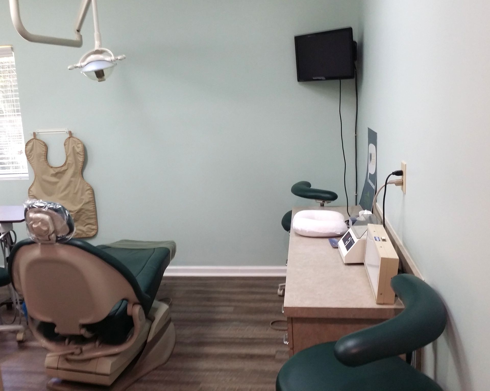 Small medical exam room with a chair, wall-mounted monitor, sink, and wooden cabinet.