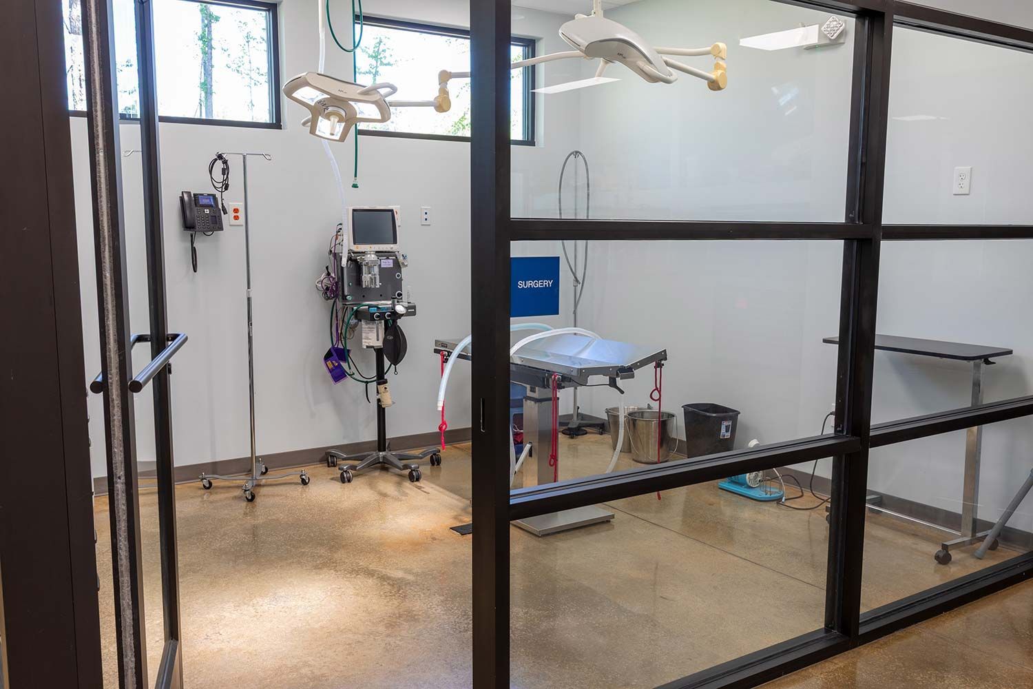 Medical exam room with table, overhead light, and equipment behind glass partition