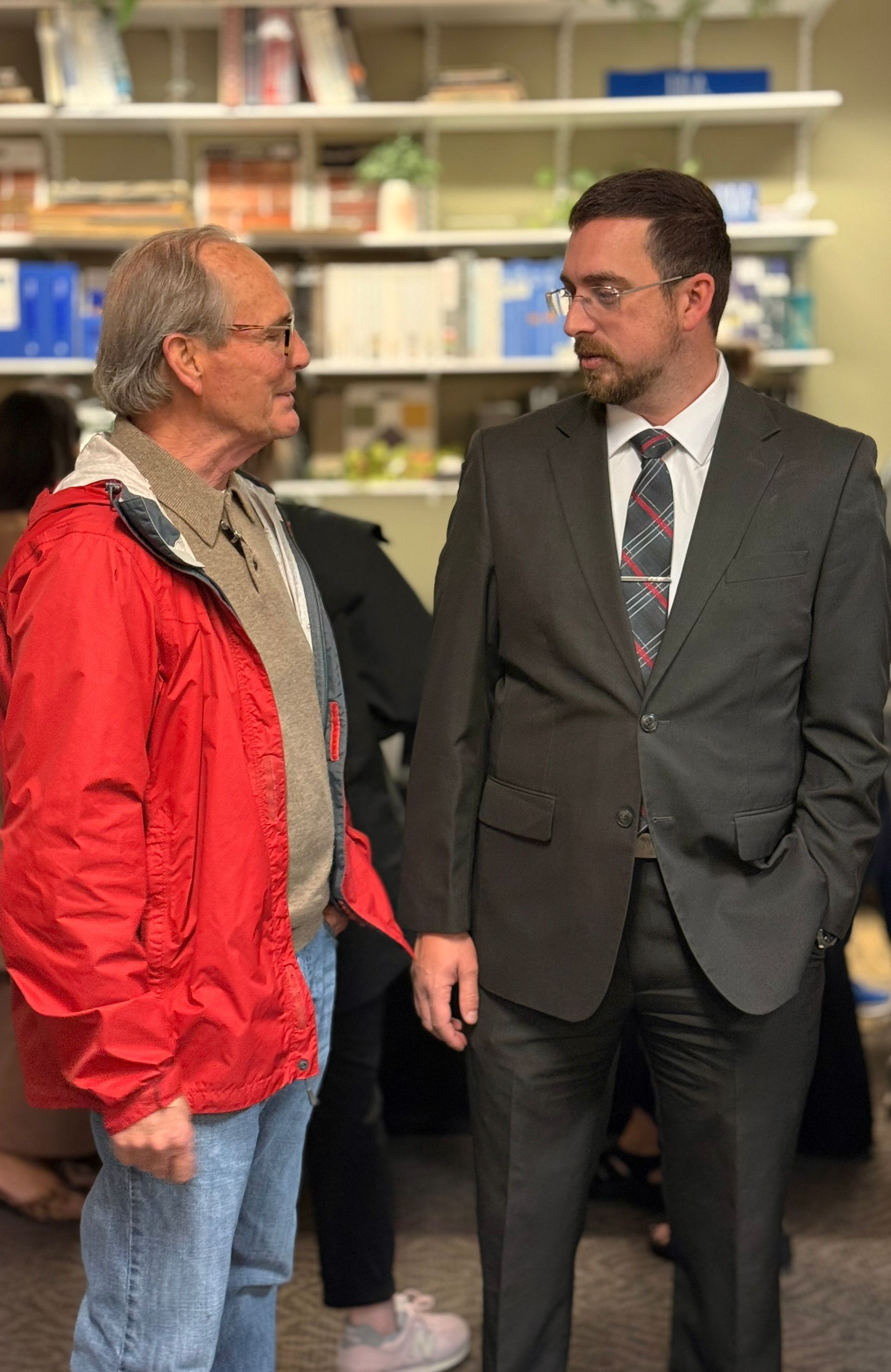 Two men talking indoors, one in a red jacket and jeans, the other in a dark suit and tie.