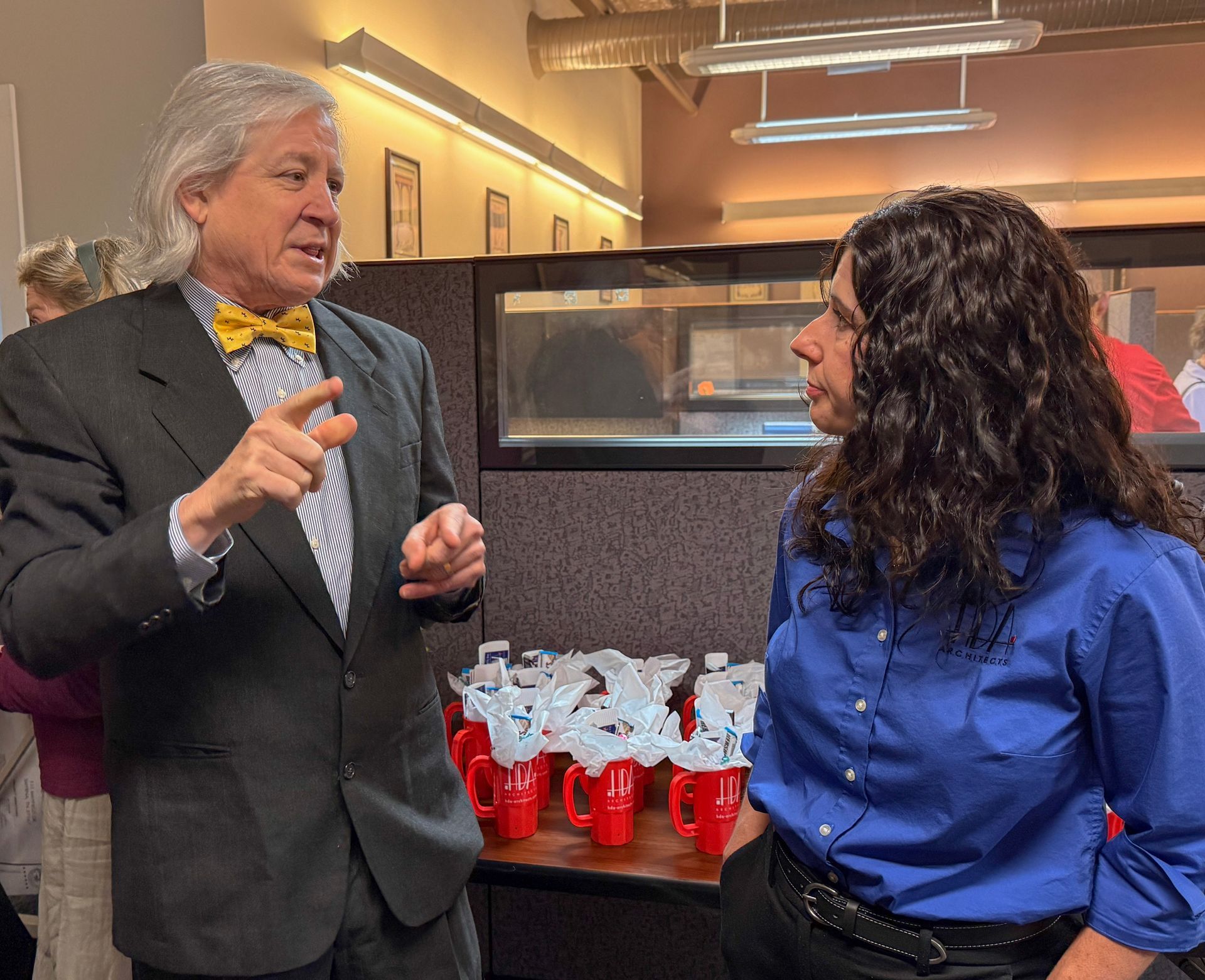 Two people talk in an office, with red mugs on a counter behind them.