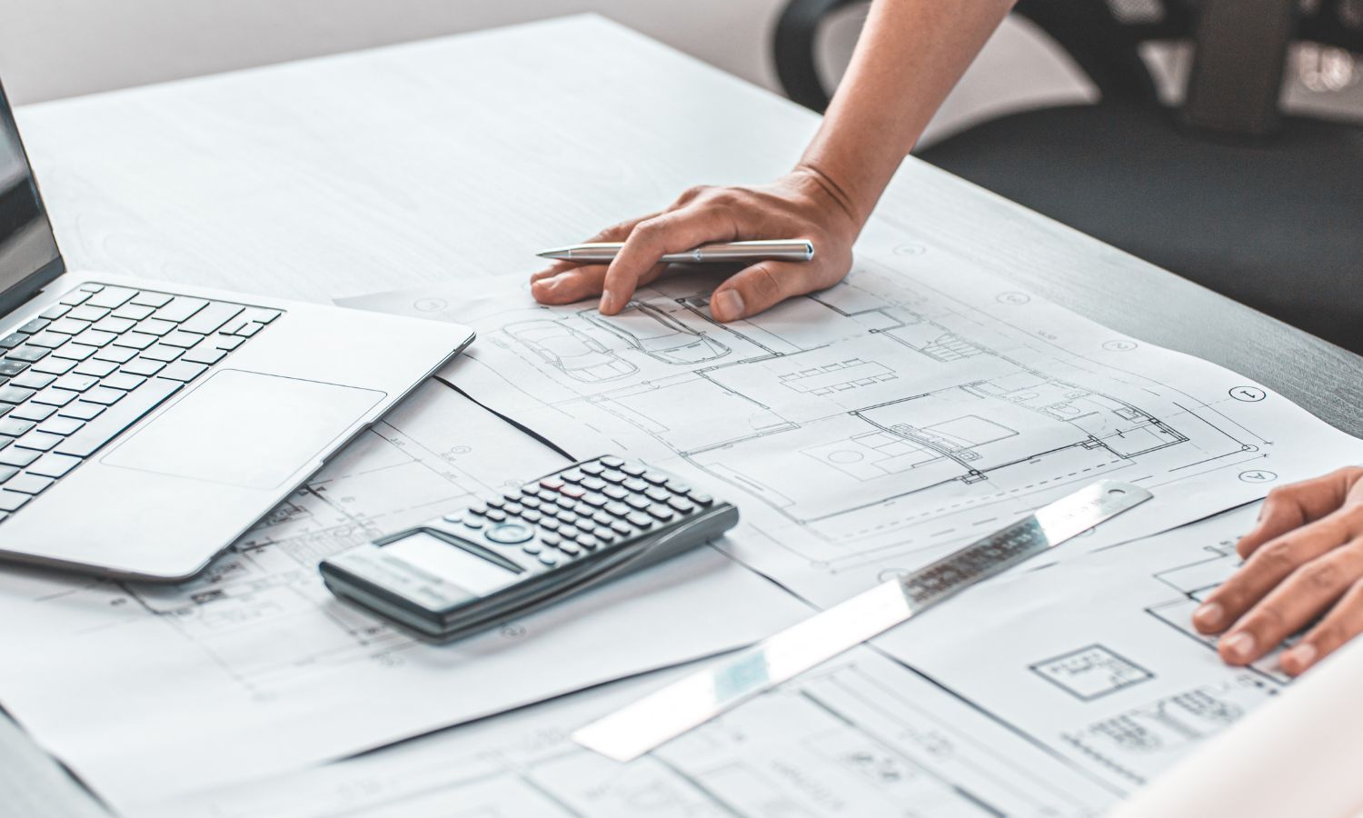Hands reviewing architectural plans with a calculator and laptop on a white desk