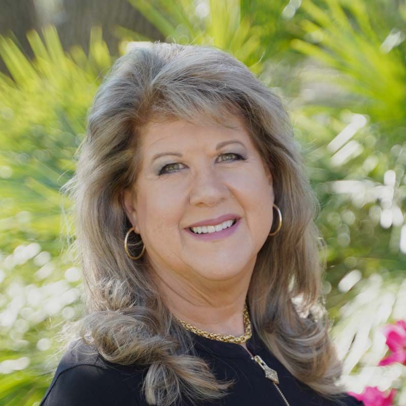 Smiling woman with gray hair and gold jewelry outdoors, with greenery in the background