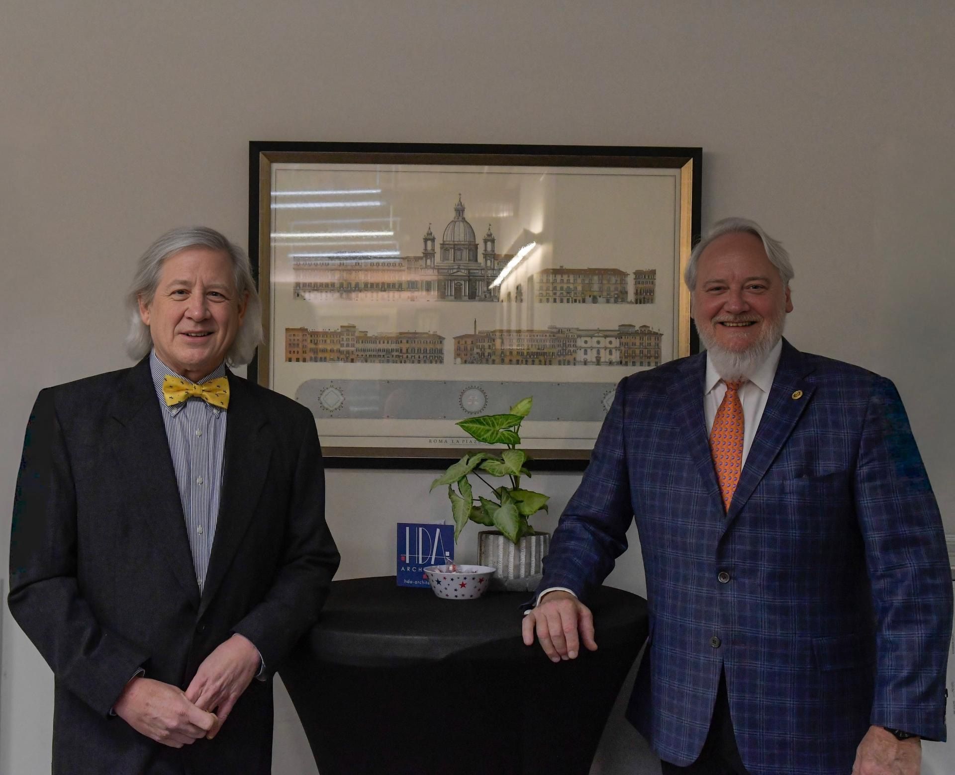 Two men in suits standing beside a table with a plant and framed artwork on the wall