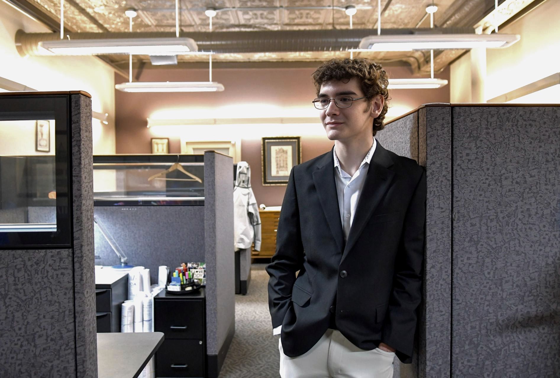 Man standing in an office cubicle area, wearing a dark blazer and white pants, looking off to the side.