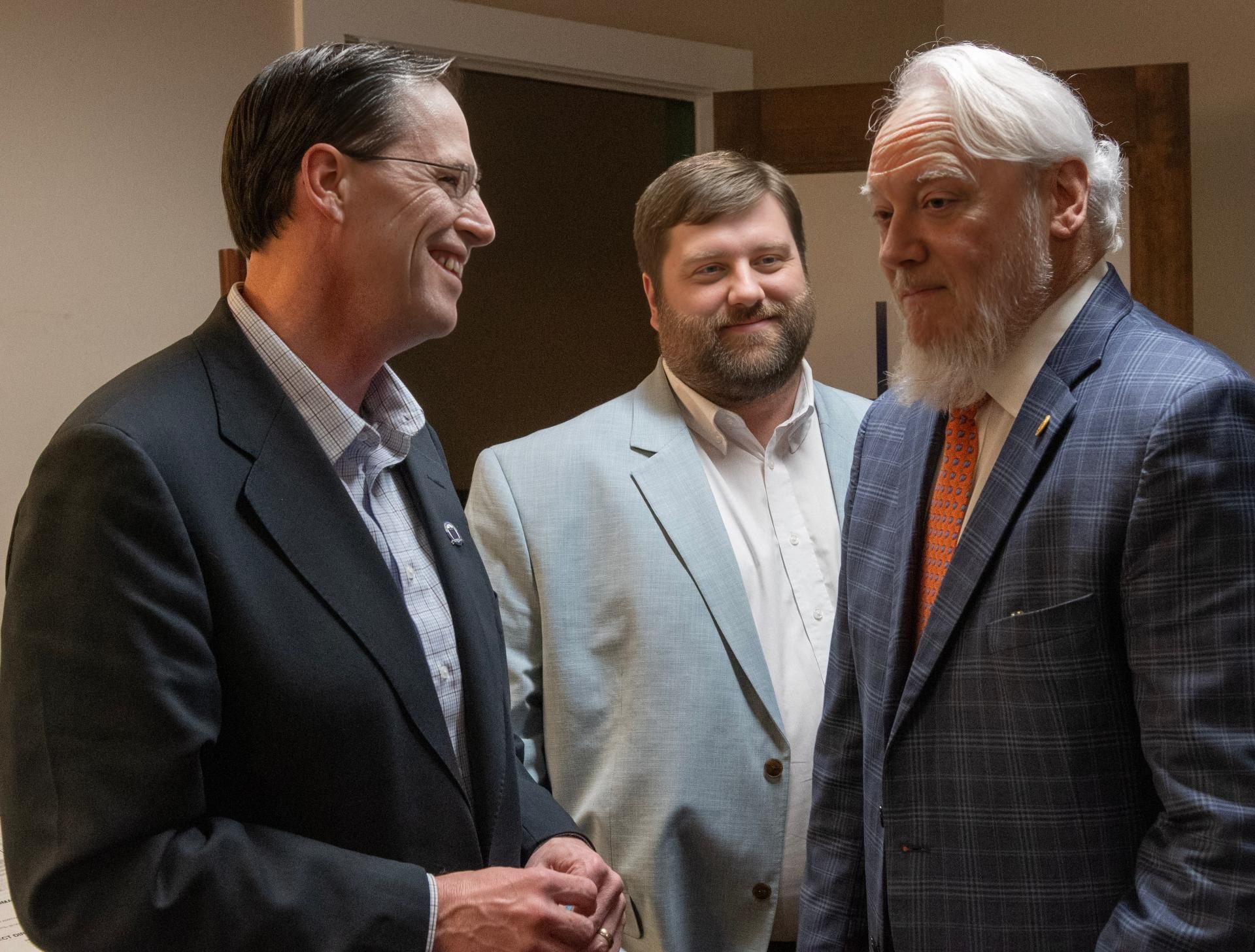 Three men in suits talking indoors, one smiling, in a warmly lit room