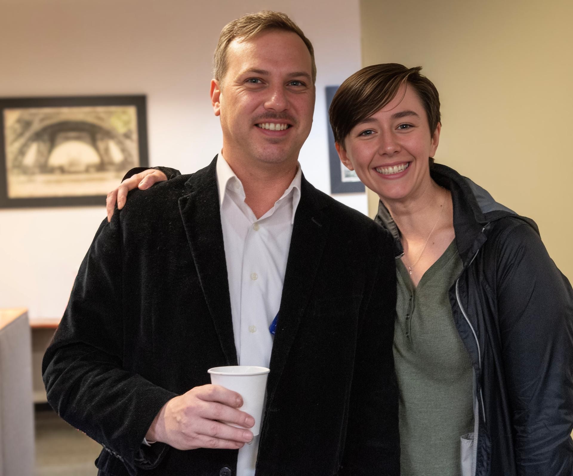 Two people smiling indoors, standing close together; one holds a white cup.