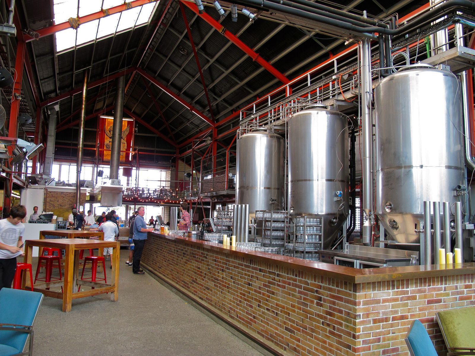 Large brewery interior with shiny stainless steel tanks and workers near a brick tasting bar