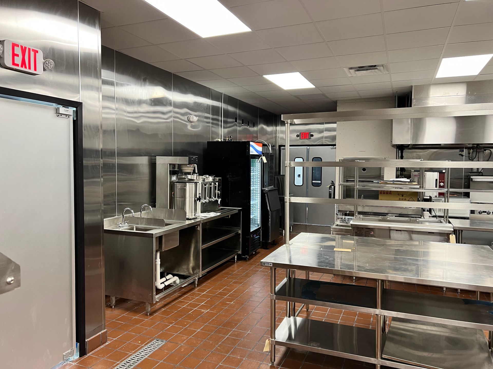 Commercial kitchen with stainless steel counters, sinks, and a black refrigerator under bright ceiling lights