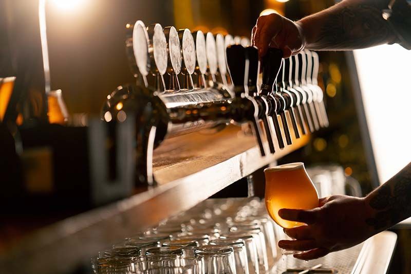 Hands pouring amber beer from taps into a glass at a bar, with rows of clean glasses below