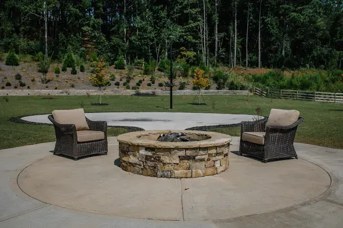 Outdoor stone fire pit with two cushioned chairs on a concrete patio, backed by grass and trees