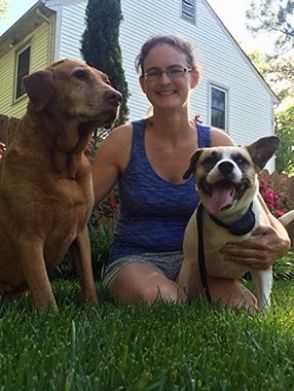 Woman sitting on grass with two dogs in front of a house, one smiling with tongue out