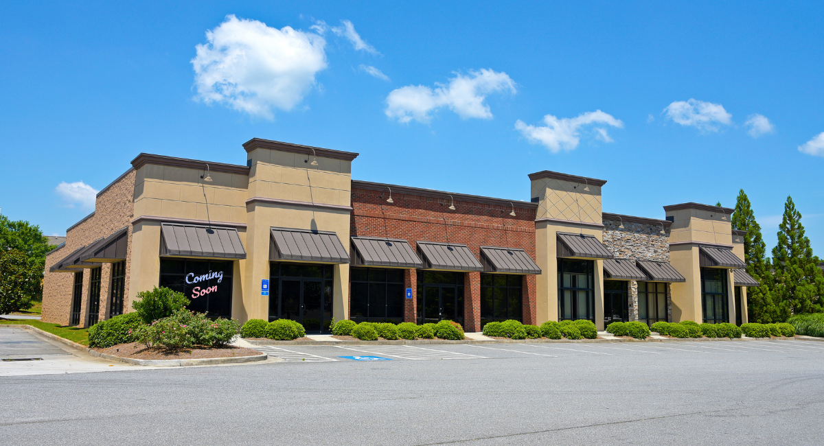 Tan stucco shopping plaza with brick accents, storefront awnings, and empty parking lot under a blue sky