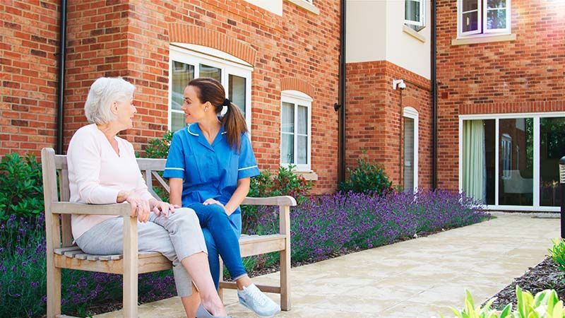 Two women sitting on a bench outside a brick building, talking in a garden courtyard