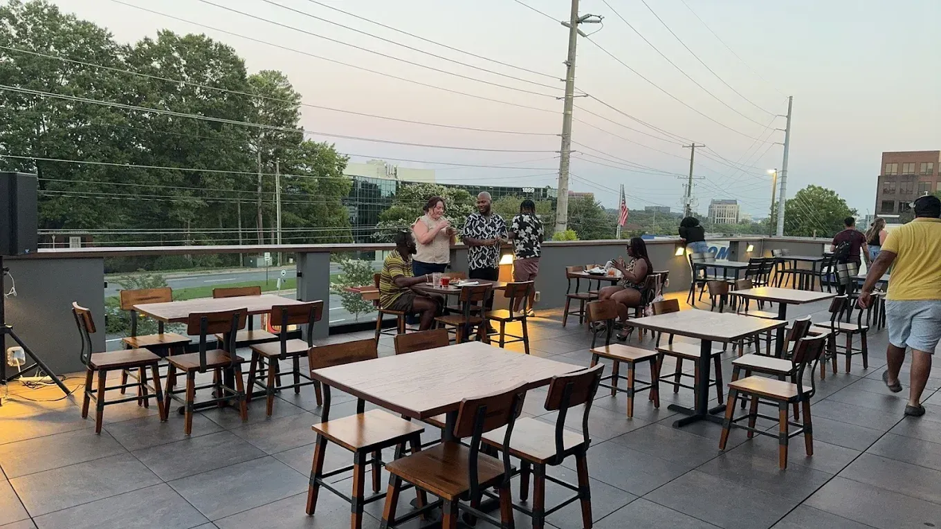 Outdoor rooftop patio with tables and chairs at dusk, a few people gathered near the railing.