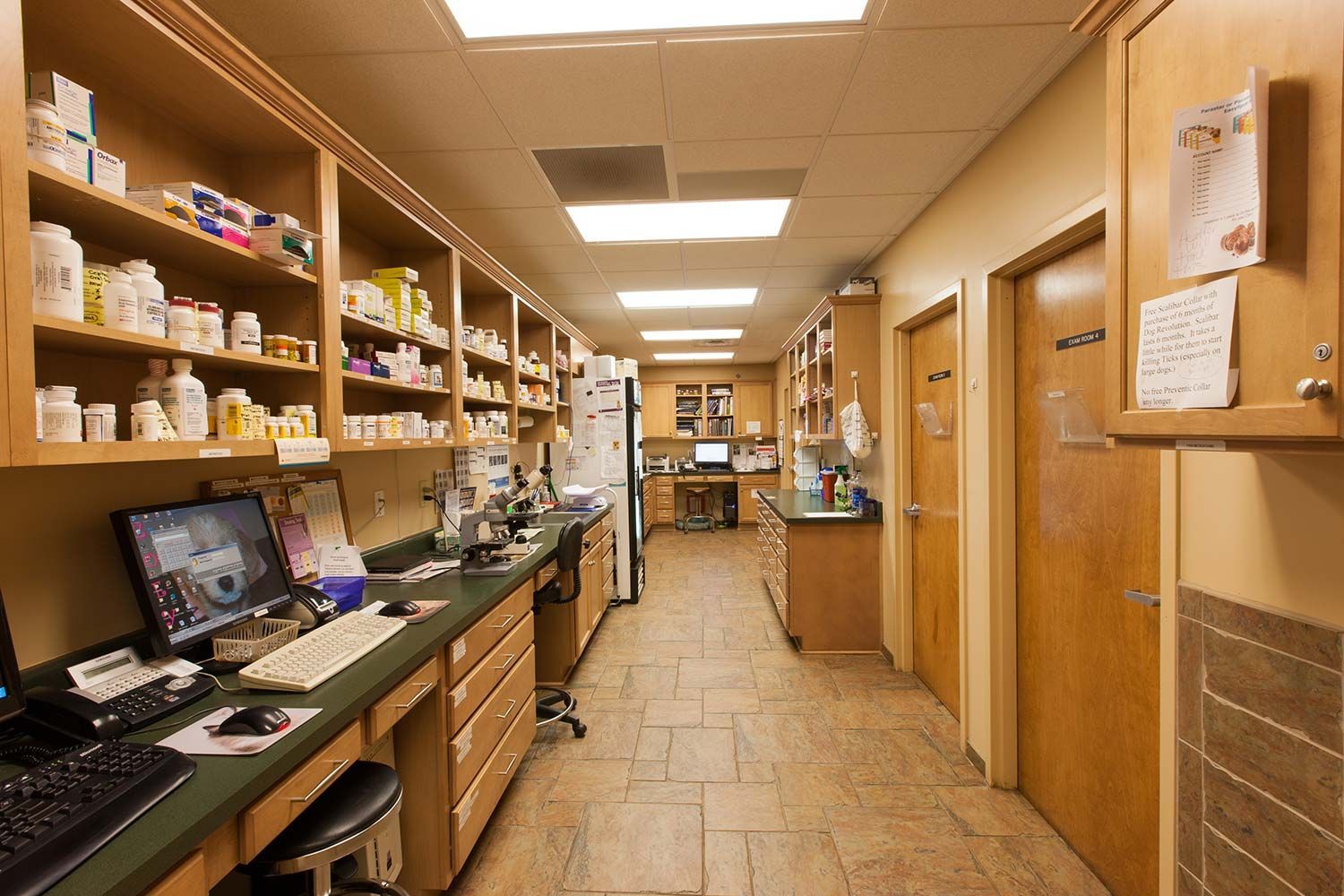 Narrow pharmacy aisle with shelves of medications, computer workstation, and wooden doors under fluorescent lights