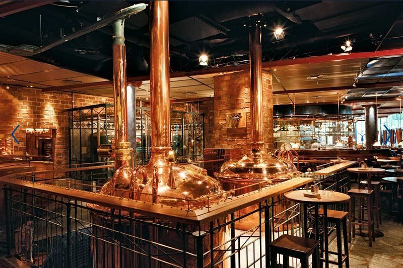 Copper brewing tanks in a dimly lit industrial-style bar or brewery with high tables and stools.