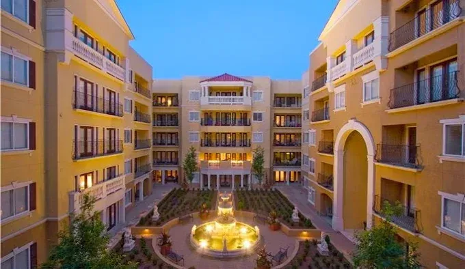 Sunlit apartment courtyard with central fountain, arched walkways, and yellow buildings around it
