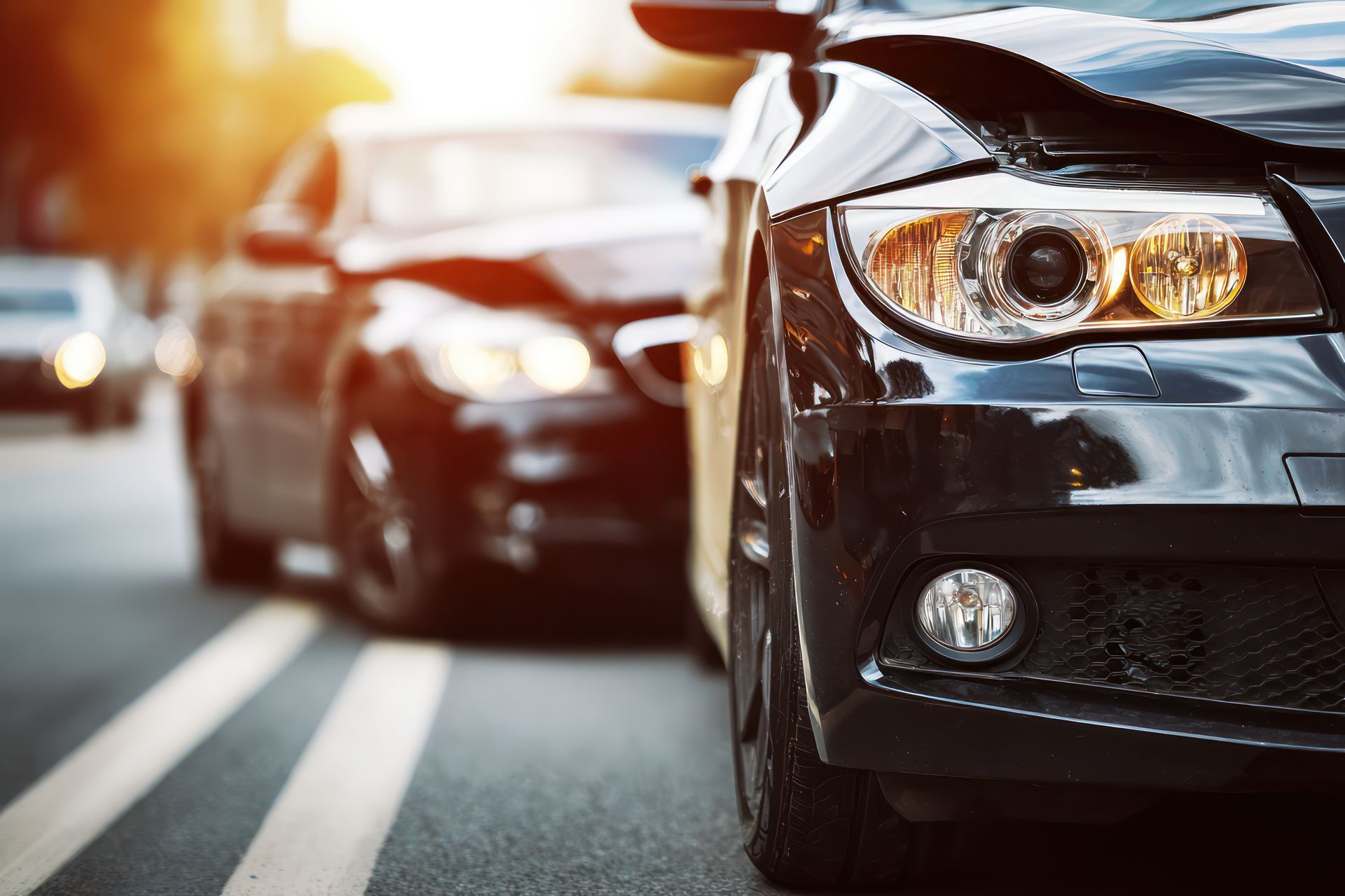 Close-up view of the front of a damaged black car, with another car partially visible in the background on a road. Close-up view of the front of a damaged black car, with another car partially visible in the background on a road.