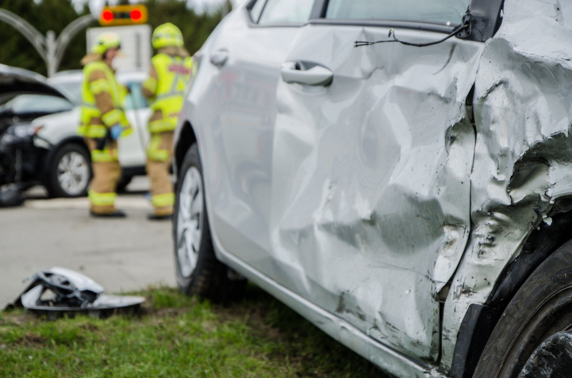 Damaged silver car side with visible dents; emergency responders in yellow vests in the background.