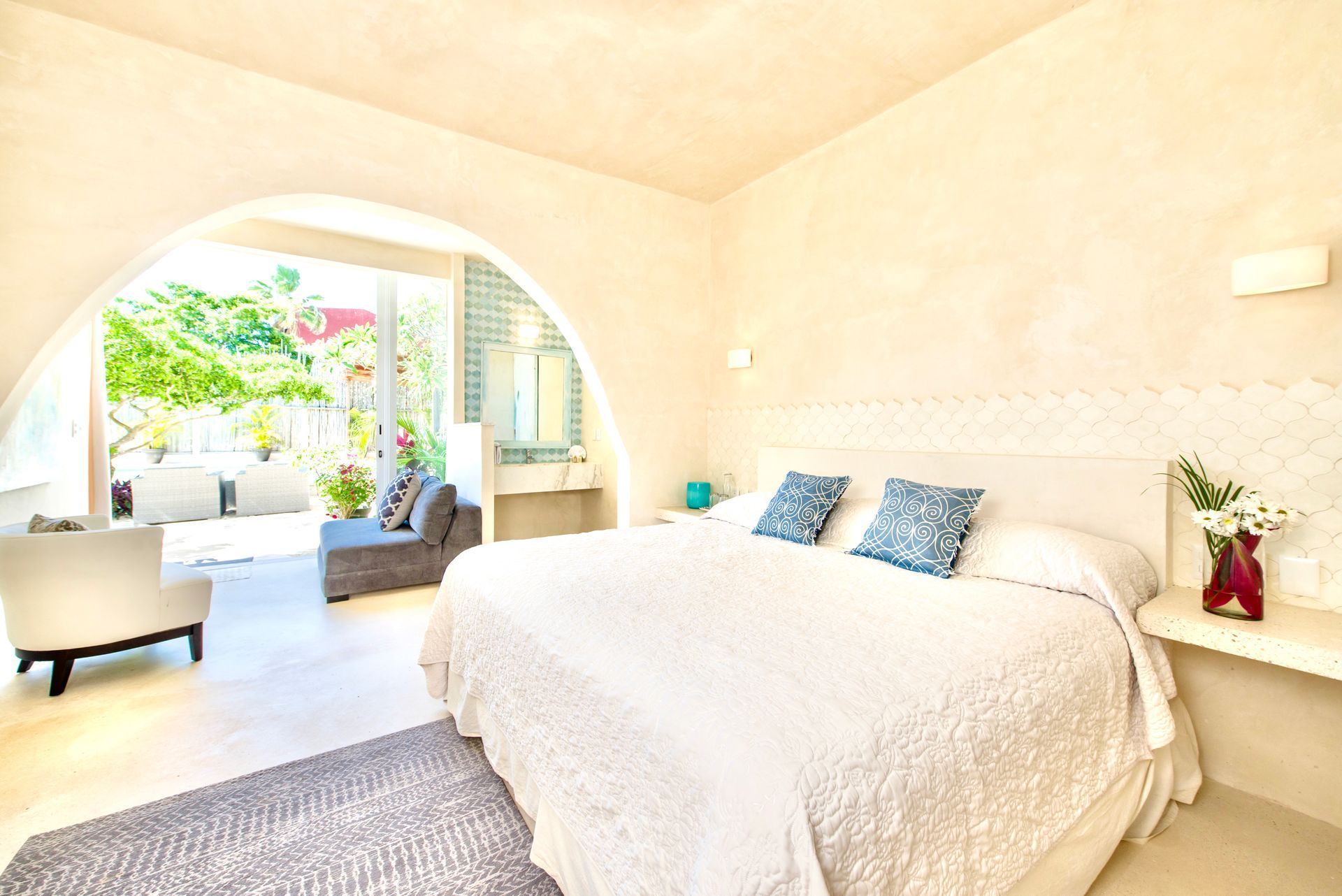 Bedroom with white walls, bed, and open archway to a patio; blue pillows, flowers.