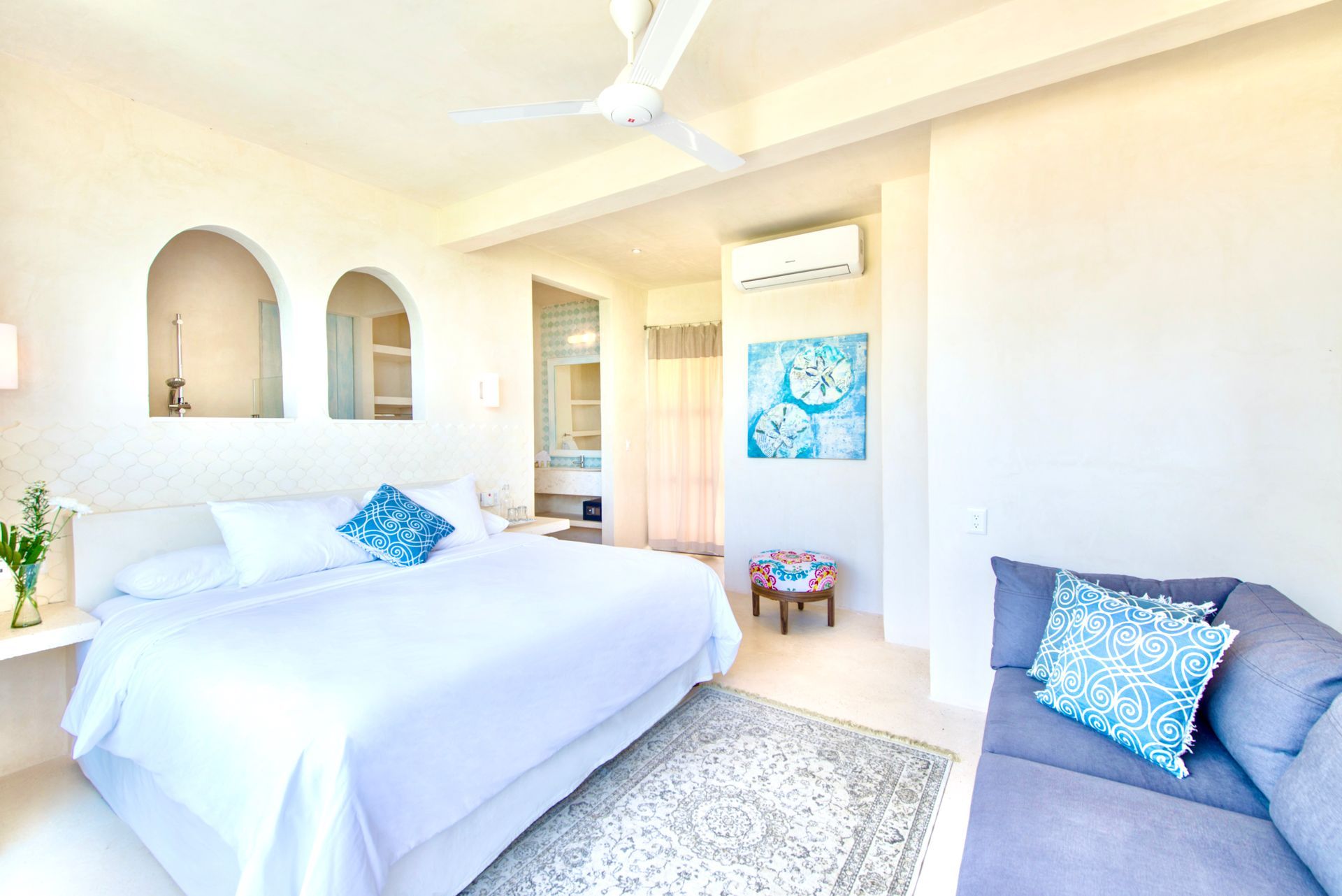 White bedroom with bed, blue accents, small sofa, artwork, and a view of the bathroom.