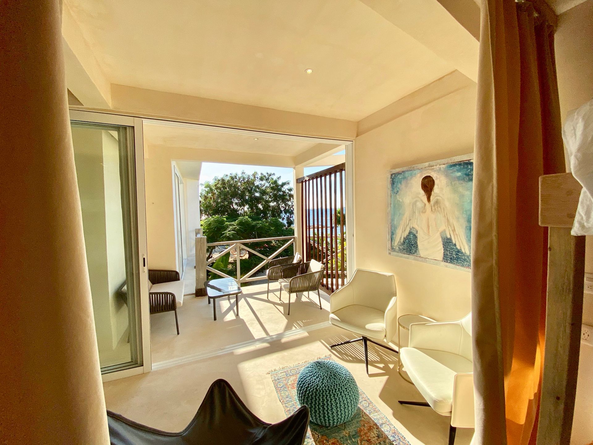 Balcony room with white chairs, table, art, and an open view to trees and the sea.