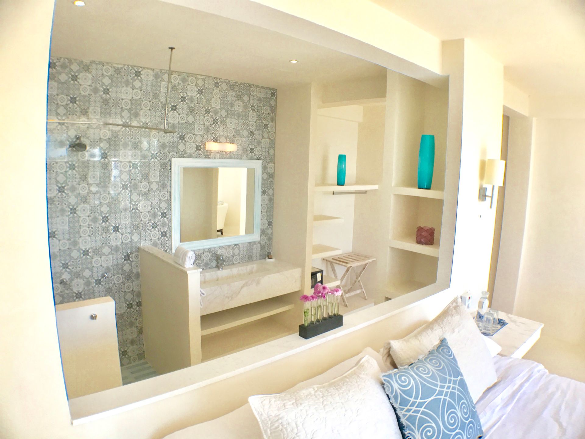 A bright bedroom featuring a bathroom with a patterned tiled wall, white vanity, and built-in shelves with teal vases.