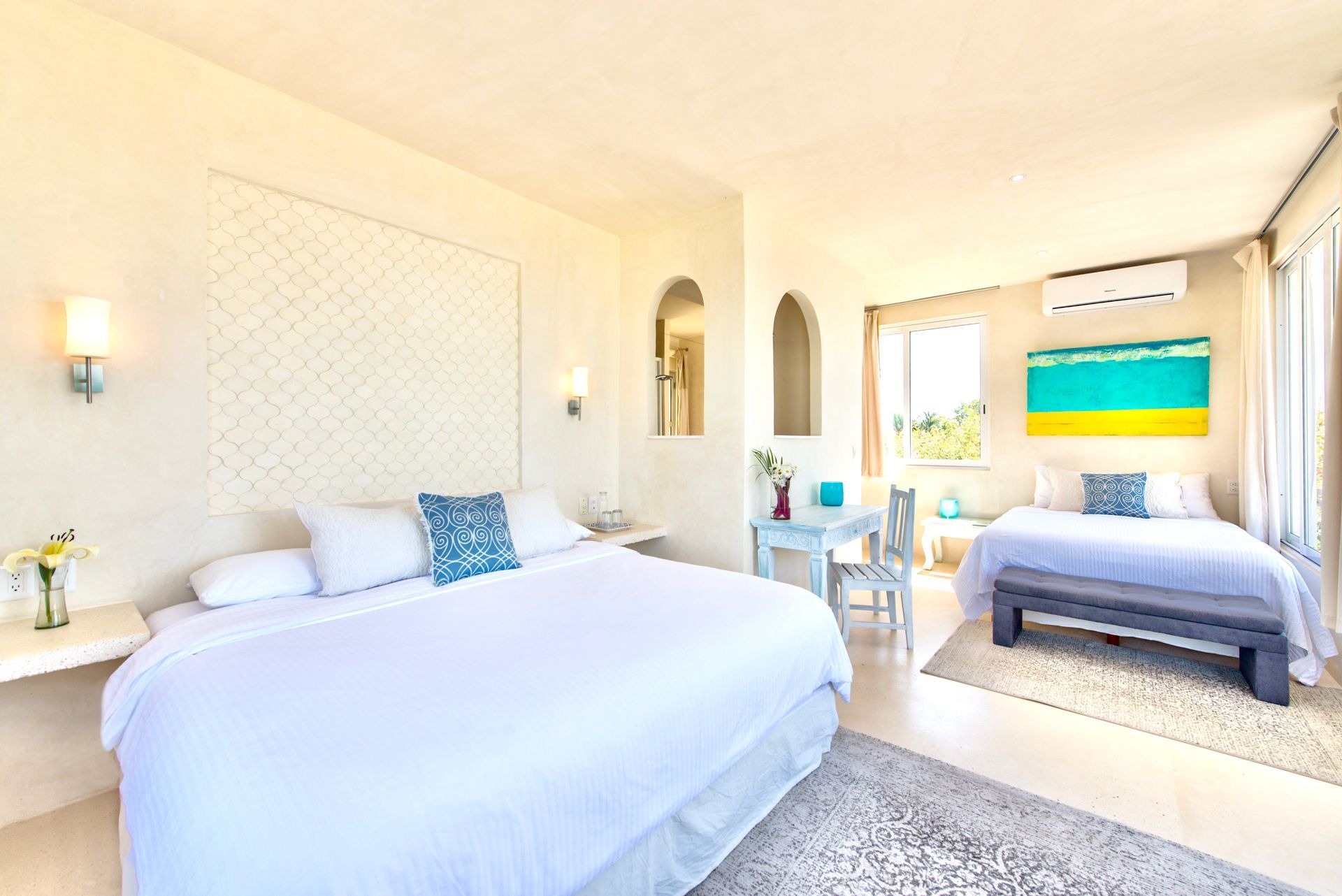 Bright bedroom with a king bed, twin bed, table, and vibrant artwork. White and blue color scheme.