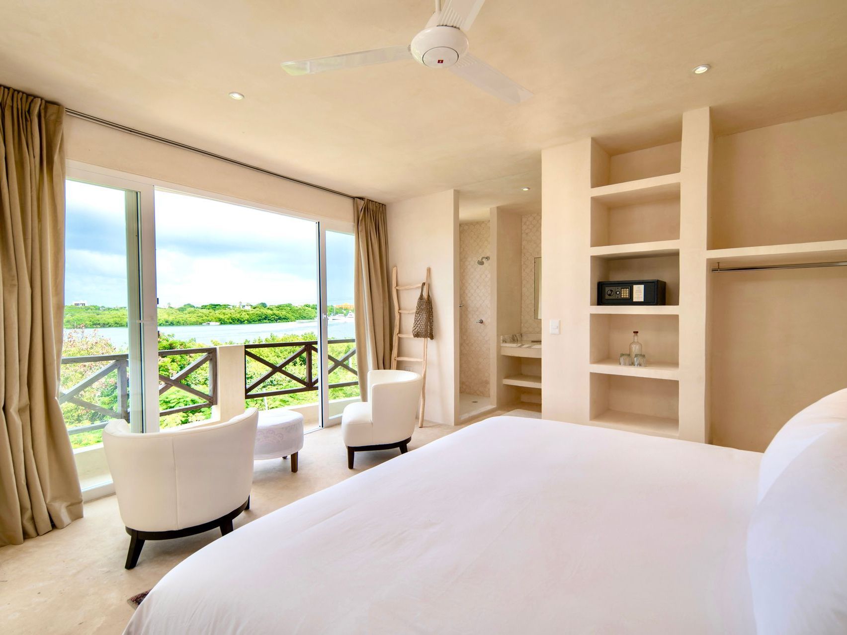 Bedroom with a large bed, two armchairs, and a balcony overlooking a body of water.