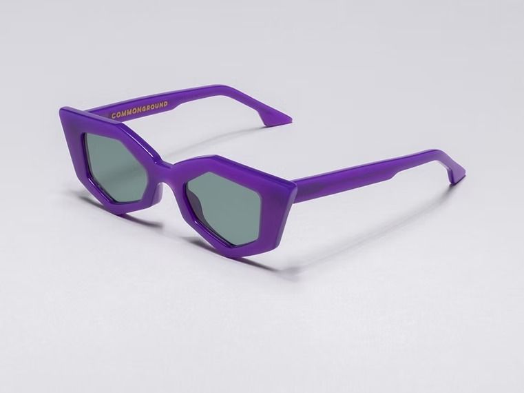 Occhiali da sole viola con montatura angolare e lenti verdi su sfondo bianco.