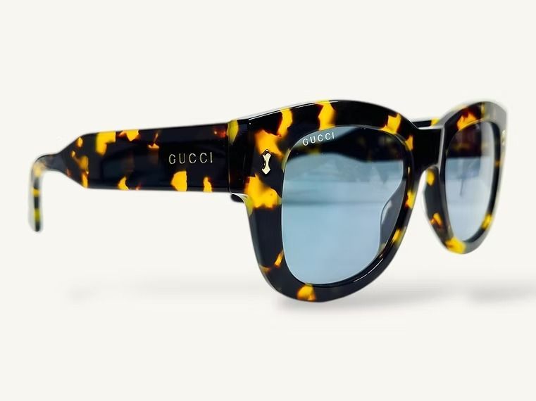 Occhiali da sole Gucci neri tartarugati con dettagli gialli e lenti blu su sfondo bianco.
