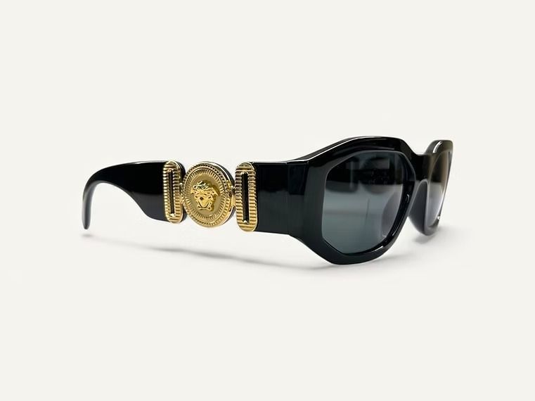 Occhiali da sole neri con medaglione decorativo dorato sulla montatura, su sfondo bianco.