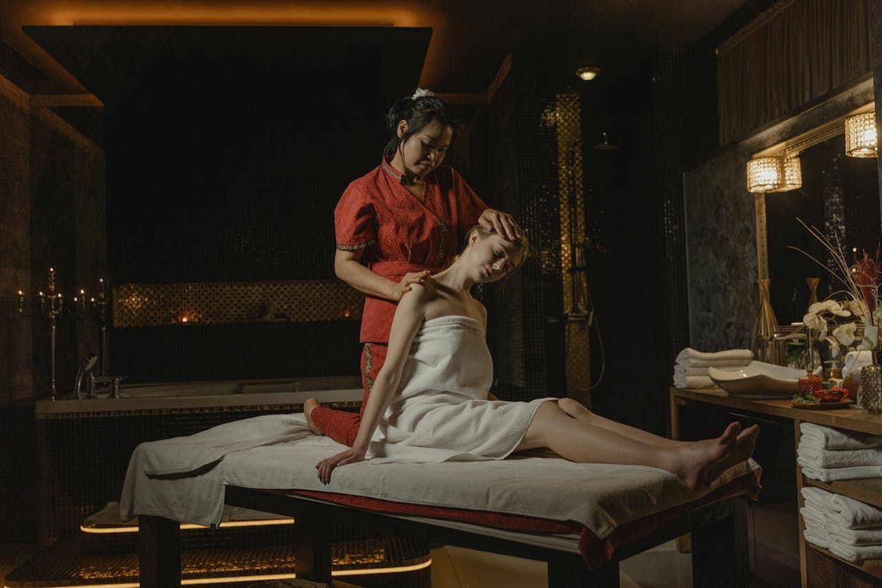 Woman receiving neck massage at a spa; masseuse in red top, spa setting, soft lighting.