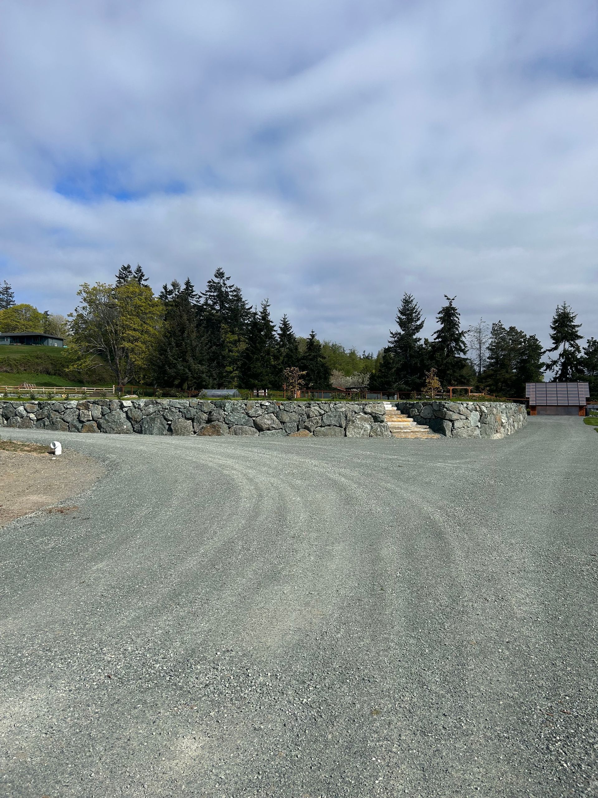 Gravel driveway leading toward a landscaped area with rock retaining walls, trees, and cloudy sky.
