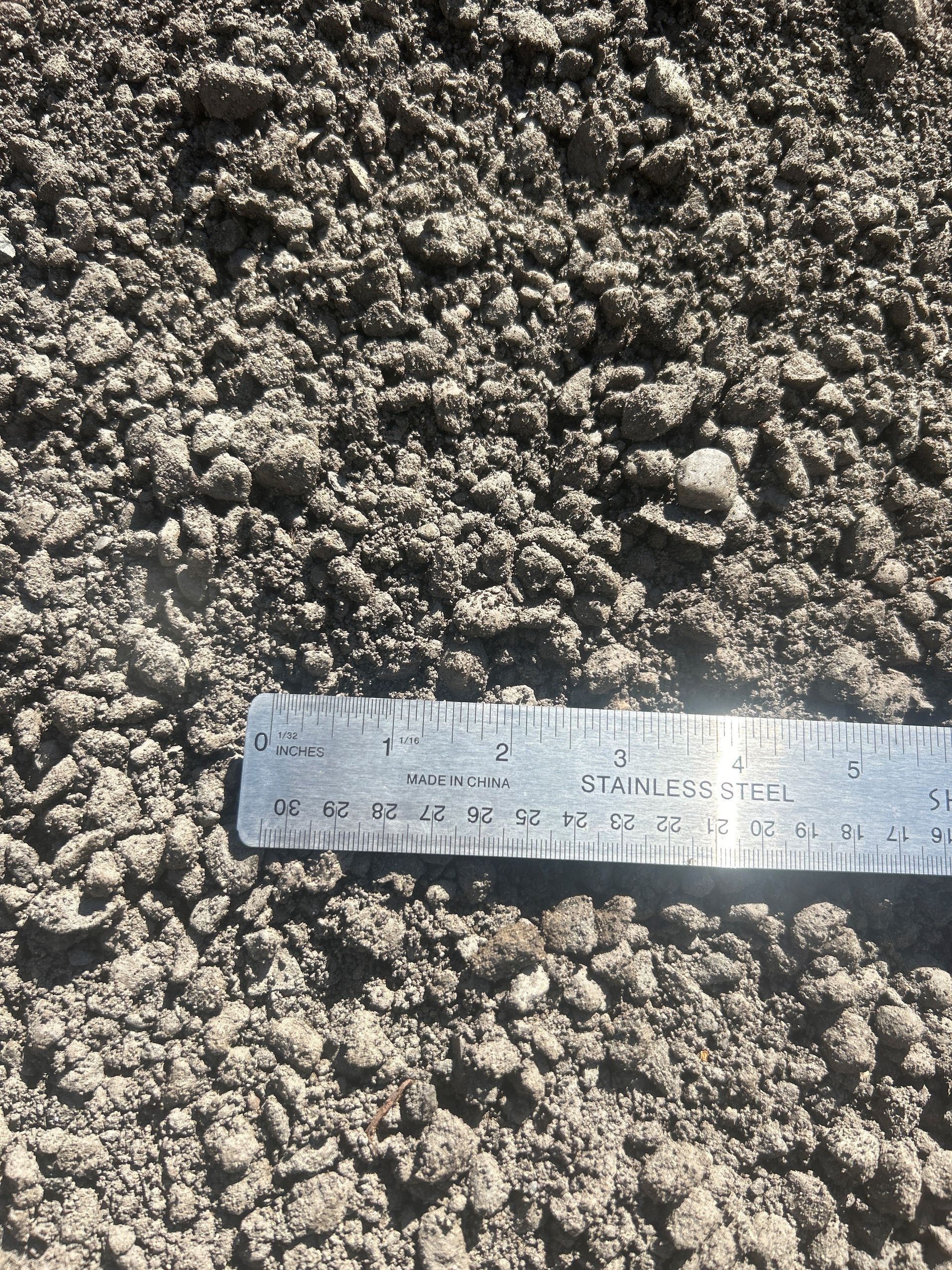 Close-up of gravel with a ruler, showing varied sizes and textures, light gray color.