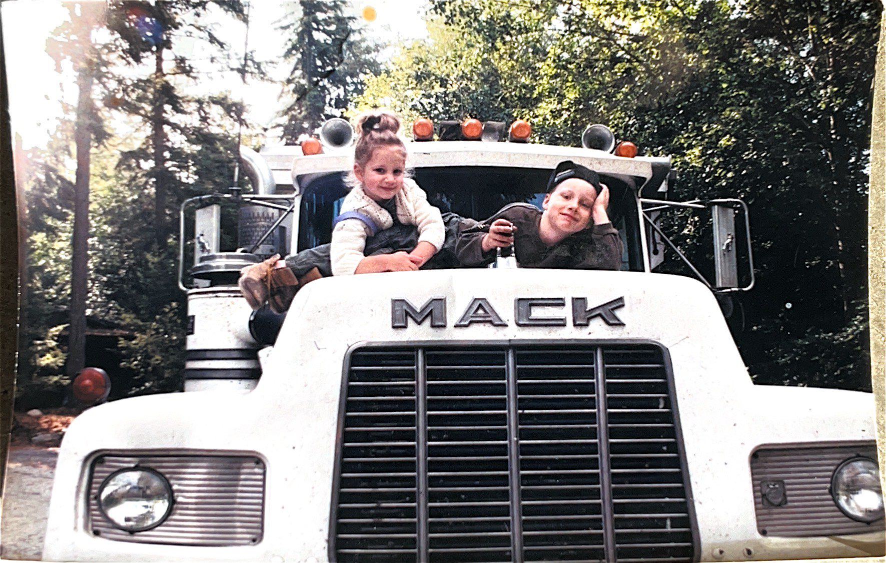 Two people smiling on top of a white Mack truck. Green trees in the background.